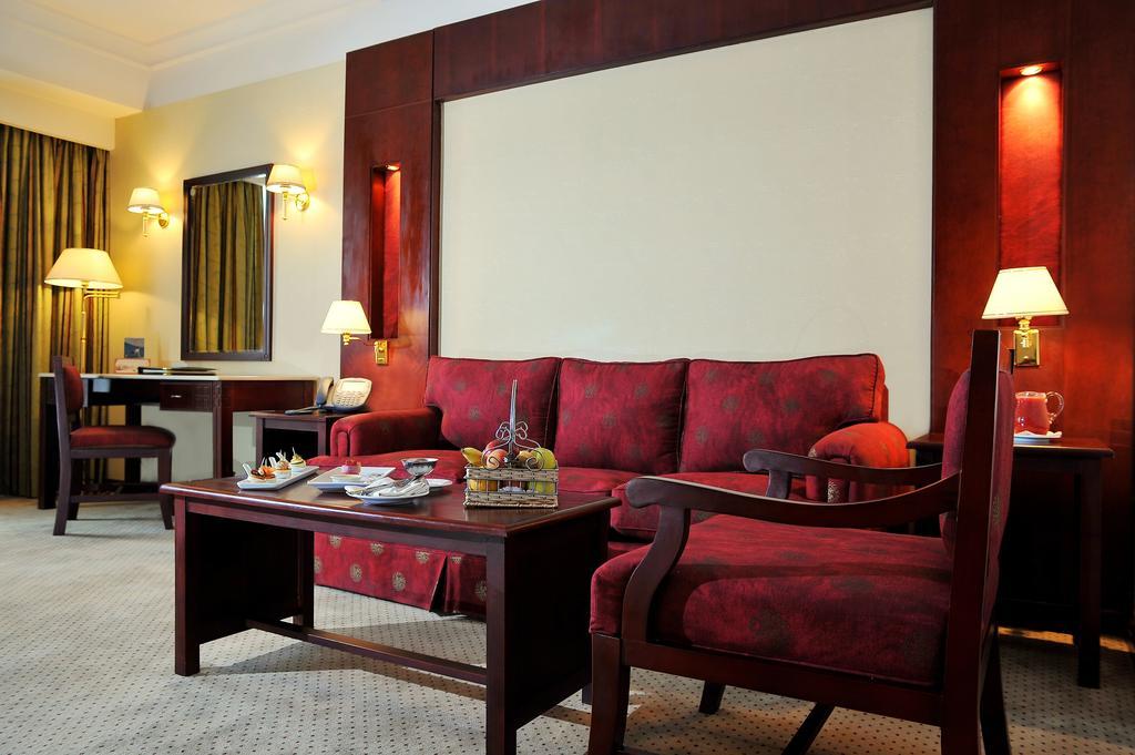 Safir Hotel Cairo Giza Phòng bức ảnh The photo shows a well-decorated hotel room or sitting area. In the center, there is a red couch with matching armchairs around it. A wooden coffee table is placed in front of the couch, and it holds some decorative items and snacks. The walls are ad