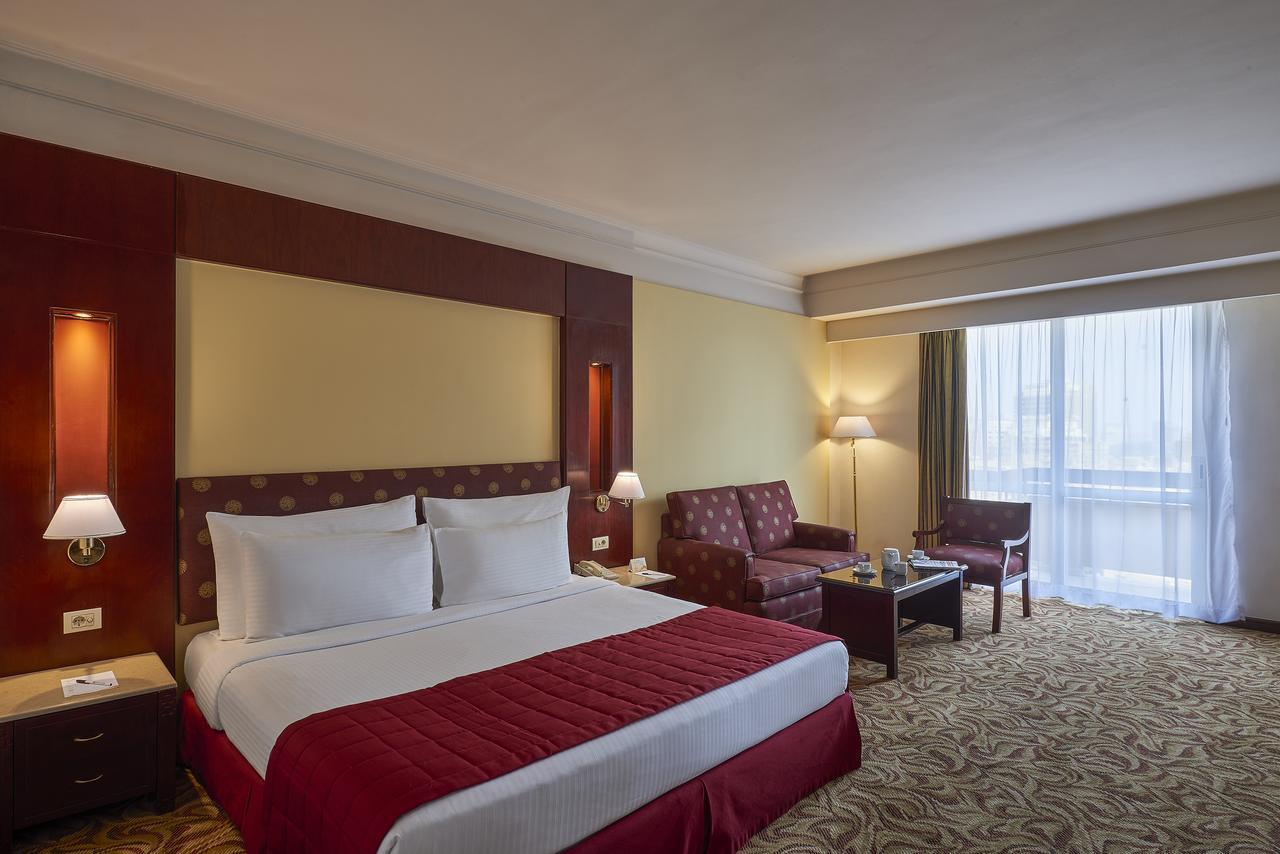 Safir Hotel Cairo Giza Ngoại thất bức ảnh The image showcases a hotel room featuring a large bed with white linens and a red bedspread. Behind the bed, there is a decorative headboard. The room includes two armchairs and a small coffee table, creating a seating area. Natural light enters thr