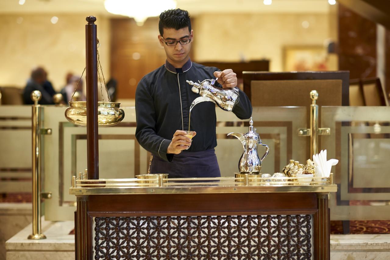 Safir Hotel Cairo Giza Ngoại thất bức ảnh The photo depicts a person at a service counter, pouring tea from a traditional teapot. The individual appears to be dressed in a formal, dark outfit, and is focused on the task of pouring. The counter features ornate decorations and various tea-serv