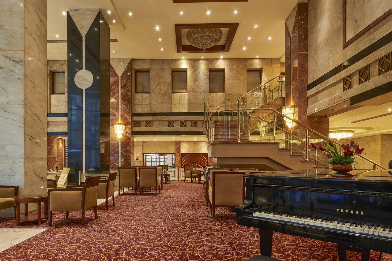 Safir Hotel Cairo Giza Ngoại thất bức ảnh The image shows an elegant hotel lobby. The space features marble walls and a plush carpet, giving it a luxurious feel. There are seating areas with comfortable chairs arranged around tables. A grand staircase leads to an upper level, and large windo