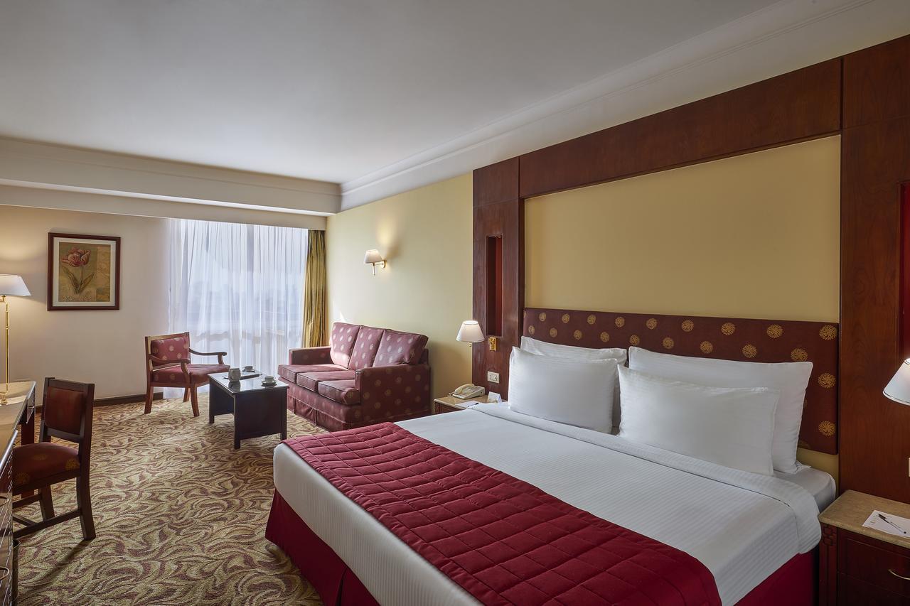 Safir Hotel Cairo Giza Ngoại thất bức ảnh The photo shows a hotel room featuring a spacious layout. In the center, there is a large bed with white bedding and a red decorative coverlet. To one side, there is a seating area that includes a sofa and two armchairs, all set around a small coffee