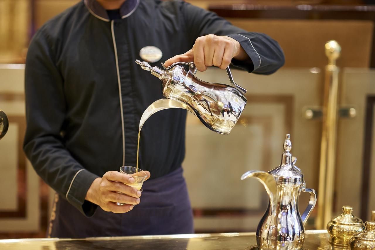 Safir Hotel Cairo Giza Ngoại thất bức ảnh The image shows a server pouring a beverage from a shiny silver teapot into a glass. The server is wearing a dark uniform and has a focused expression. The teapot is intricately designed, and there is another similar pot alongside it on the counter. 