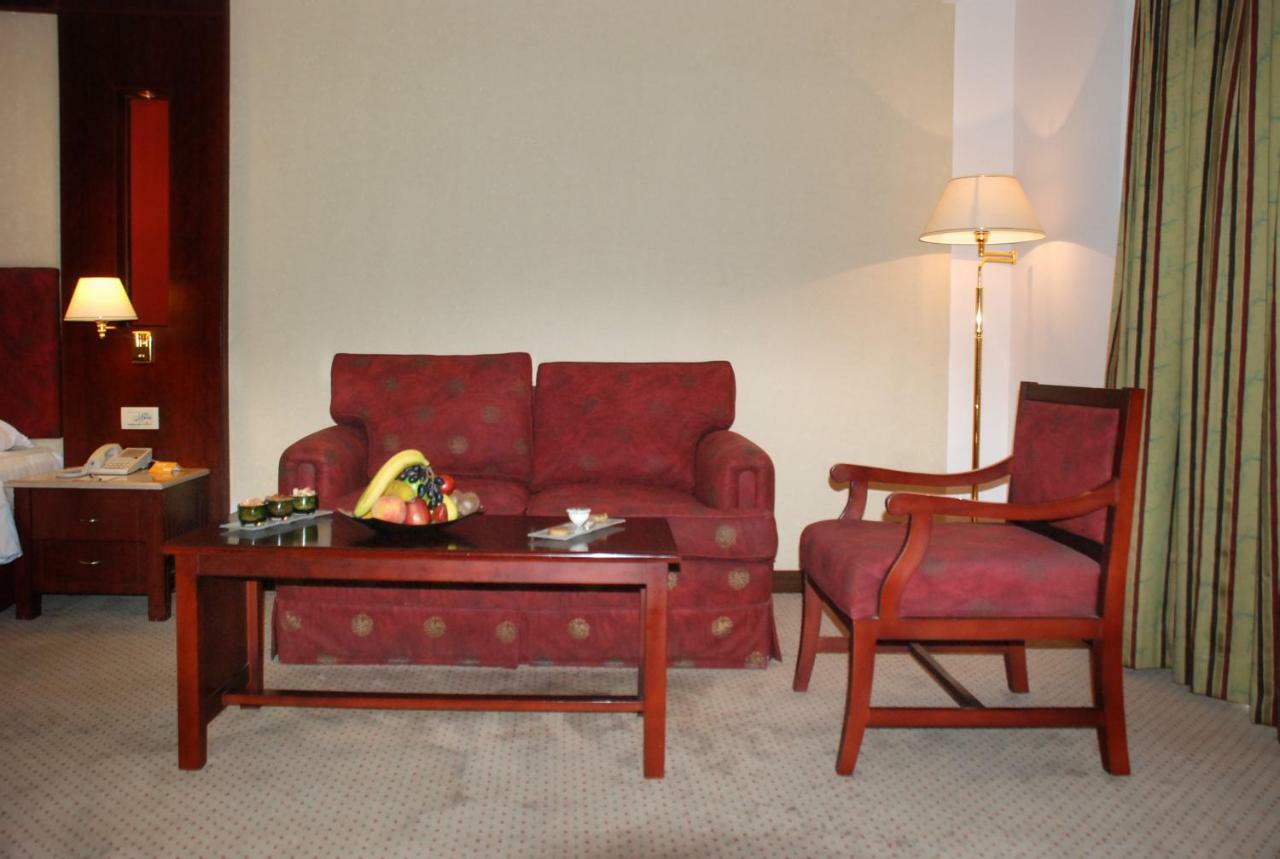 Safir Hotel Cairo Giza Ngoại thất bức ảnh The photo shows a cozy seating area in a hotel room. There is a red patterned sofa and a matching armchair placed around a wooden coffee table. On the table, there are some snacks or fruits displayed. A floor lamp is positioned beside the sofa, and t