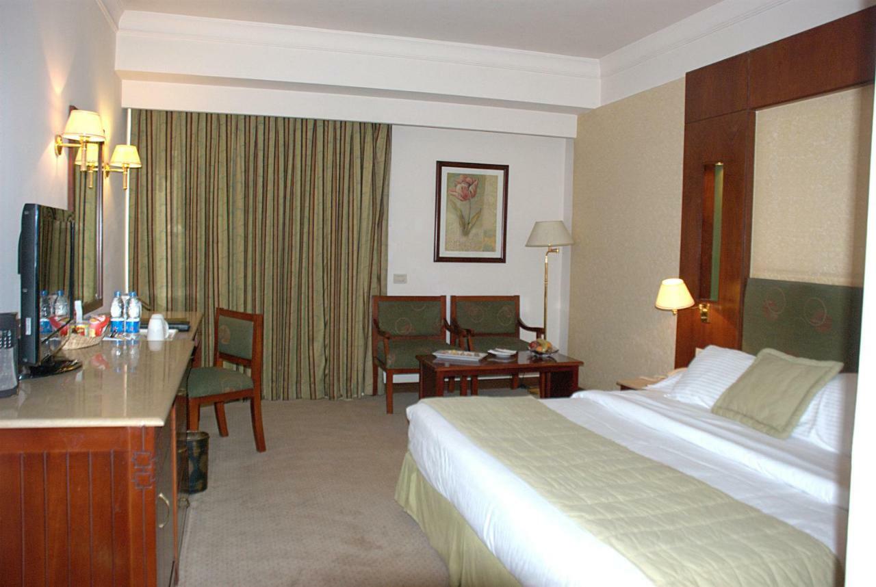 Safir Hotel Cairo Giza Phòng bức ảnh The photo shows a hotel room. It features a bed with white linens and a green throw. On one side of the bed, there's a small bedside lamp. Opposite the bed is a desk with a mirror above it, alongside a couple of chairs and a small table. The room has
