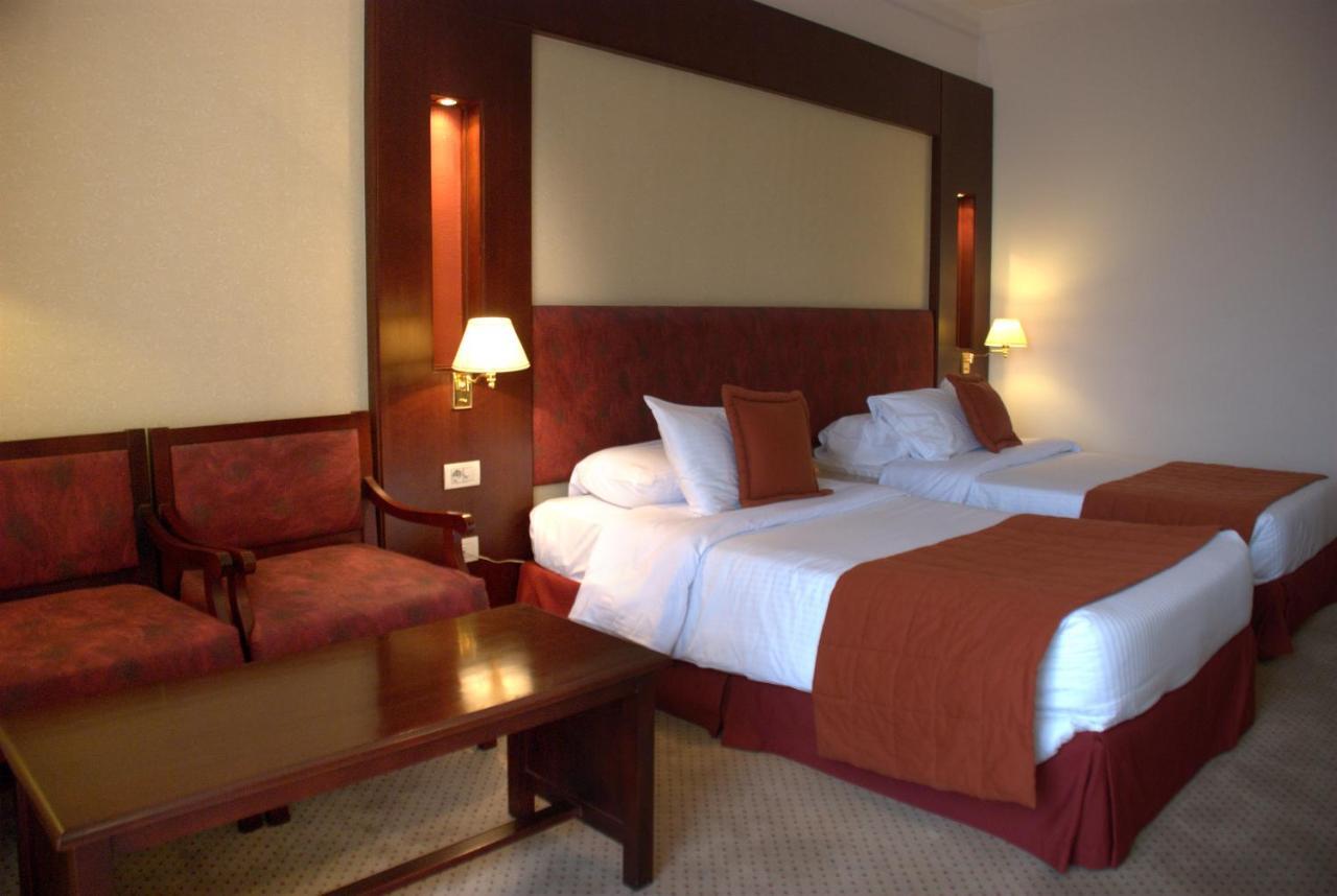 Safir Hotel Cairo Giza Ngoại thất bức ảnh The photo shows a hotel room featuring two twin beds with white linens and orange bedspreads. There is a headboard behind the beds, along with wall-mounted lamps on either side. In the corner, there are two upholstered chairs and a wooden coffee tabl